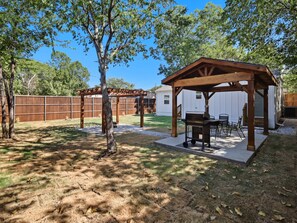 Patio trasero con una pérgola de madera, una parrilla y mobiliario para exterior.