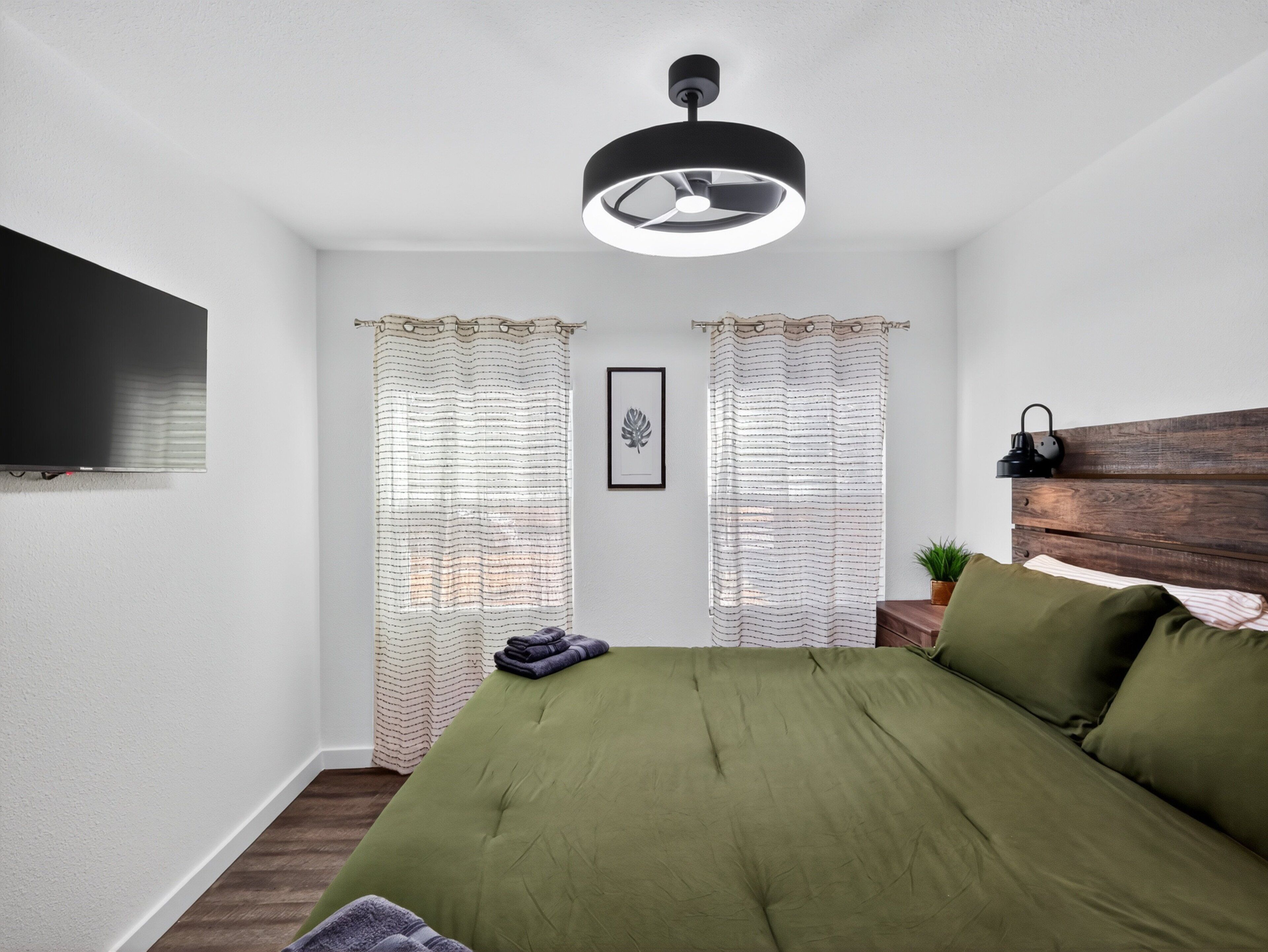 A bedroom with a bed, a flat-screen TV, a ceiling fan, and two windows with curtains.