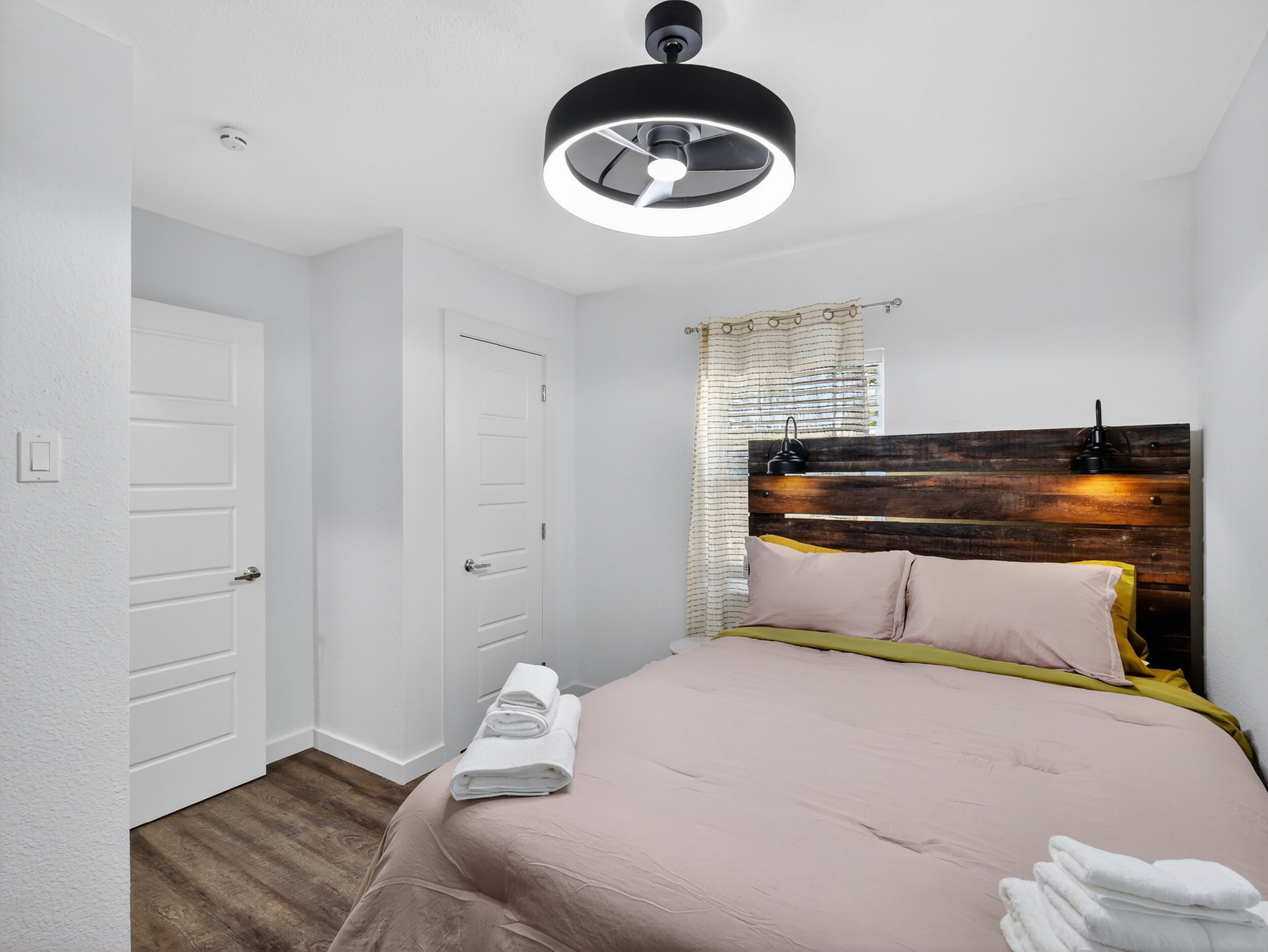 Een slaapkamer met een houten hoofdbord, een bed met roze beddengoed, een raam met gordijnen en een plafondlamp.