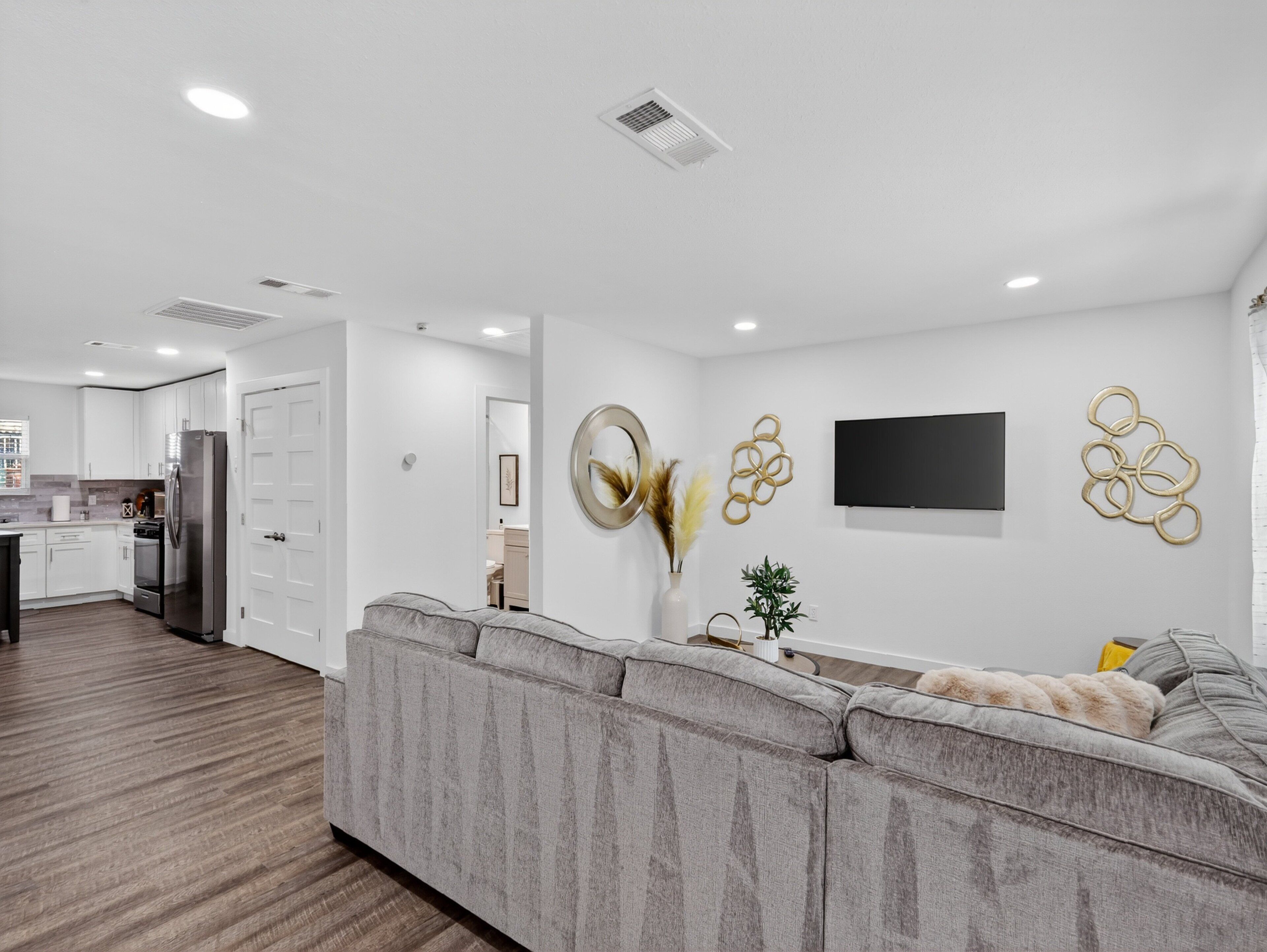 A modern living room with a grey sectional sofa, a flat-screen TV, decorative wall art, and a kitchen in the background.