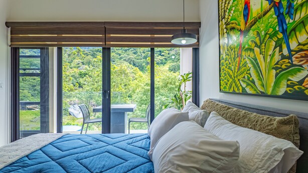 Un dormitorio con una cama grande, vistas a un jardín exuberante a través de una puerta corrediza de vidrio, y un cuadro de follaje tropical en la pared.