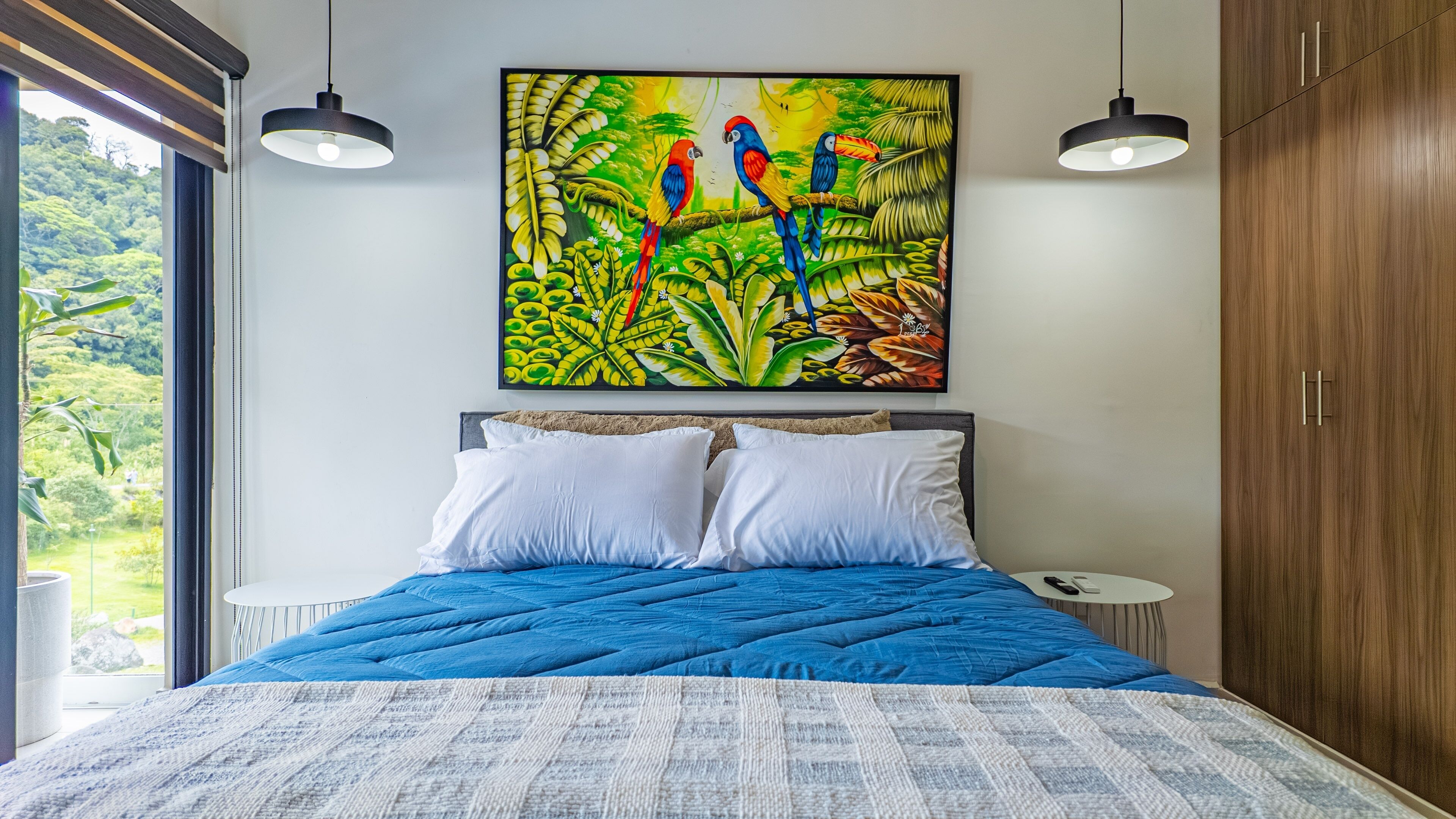 Een slaapkamer met een groot bed, twee witte kussens, een blauwe quilt, een schilderij van kleurrijke papegaaien, en een houten garderobe.