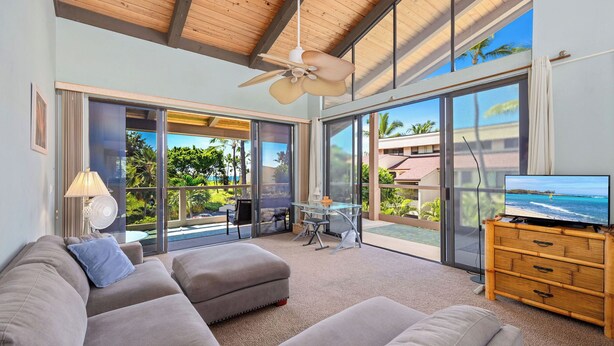 A modern living room with a large sofa, a television on a wooden cabinet, and a balcony with a view of the ocean. - Kksr#6301 Ocean/Golf Course Views, Top Floor, Corner Unit, Cathedral Ceilings (Kailua Kona)