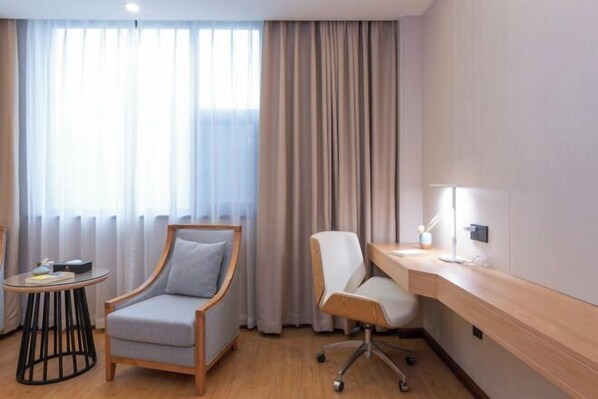 A hotel room with a wooden desk, two chairs, a small side table, and a large window with curtains.