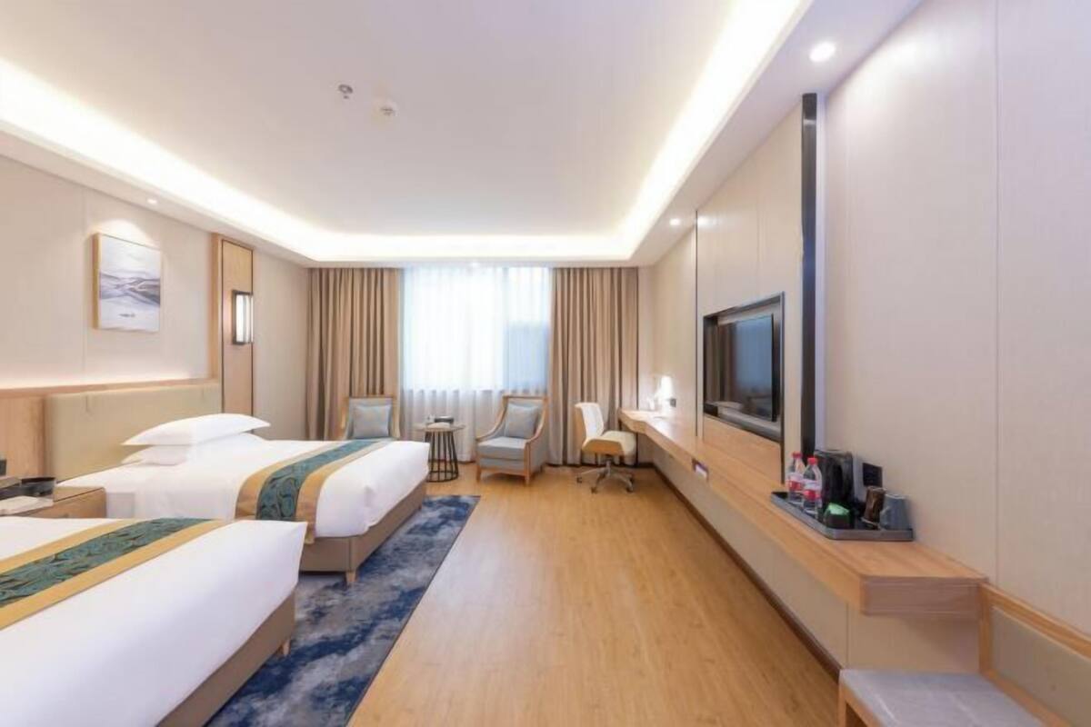 A hotel room with two beds, a desk, a chair, a TV, and a small table with items on it.