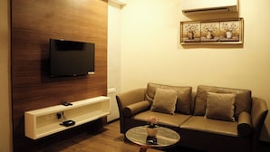 A modern living room with a flat-screen TV mounted on a wooden wall, a leather sofa with cushions, a small round coffee table with a plant, and a white entertainment unit below the TV. - The Platinum Boutique Inn Hotel (Hyderabad)
