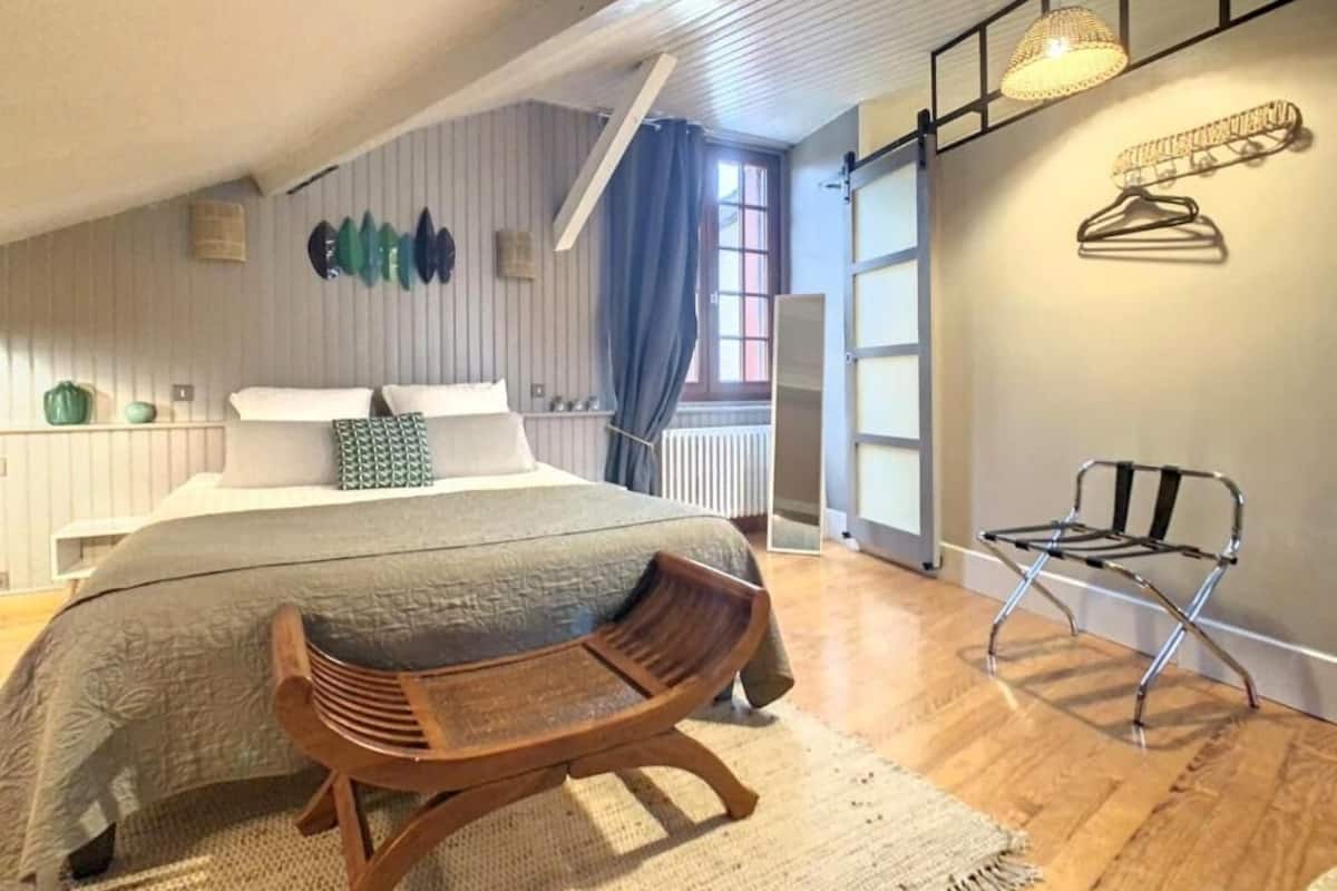 Une chambre à coucher avec un lit, une chaise en bois, un luminaire suspendu et une fenêtre avec des rideaux.
