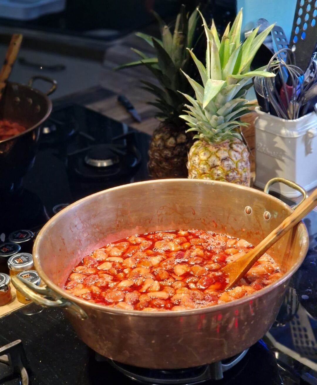 A large pot on a stove with a wooden spoon inside, surrounded by kitchen utensils and a pineapple.
