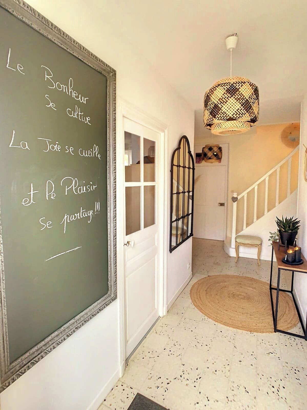 Un couloir avec un tableau noir où sont écrites des expressions françaises, un escalier, un luminaire en fibres tressées et une petite table sur laquelle est posée une plante.