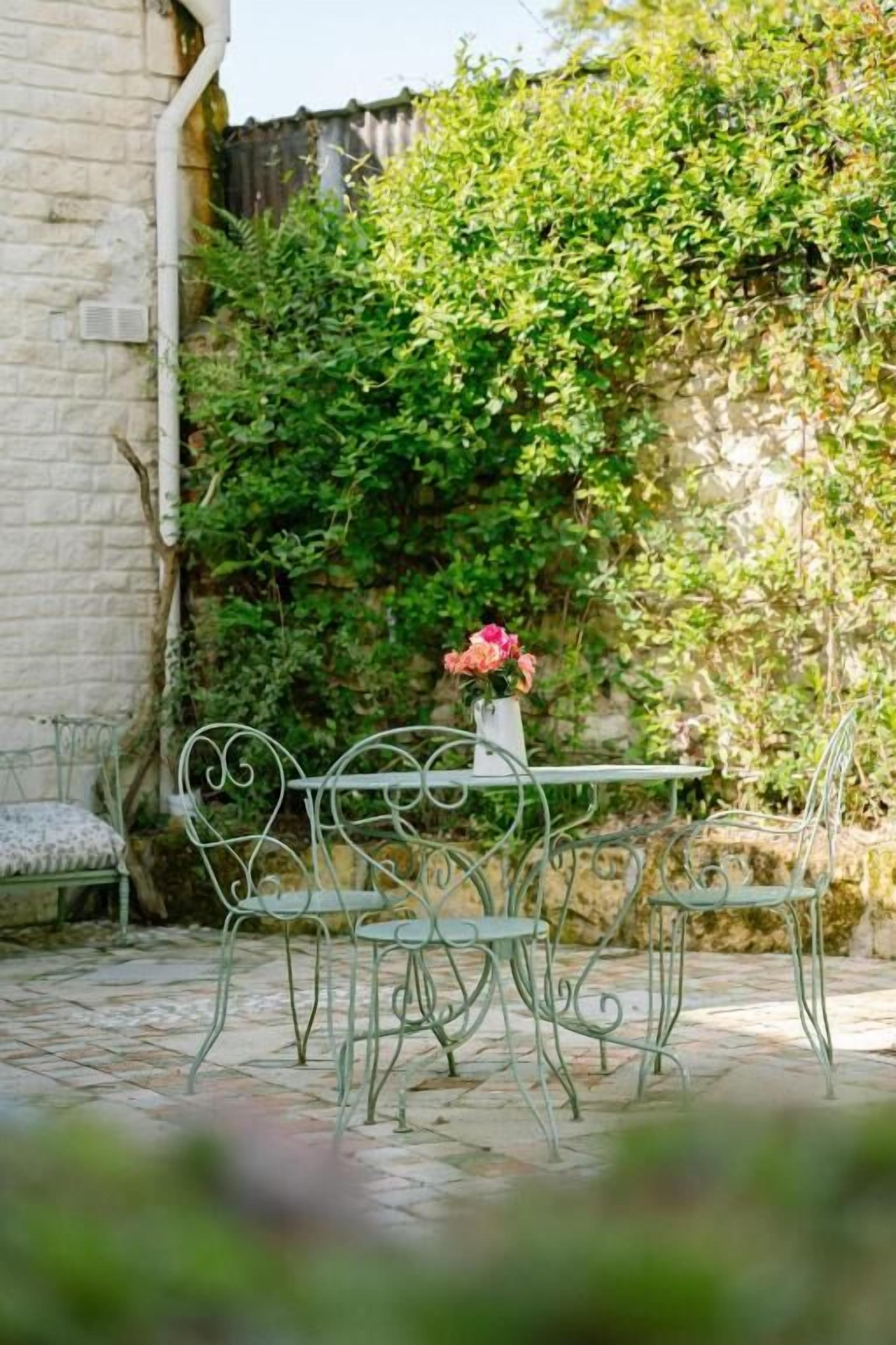 Un patio al aire libre con una mesa y sillas de metal, un banco y un jarrón con flores.