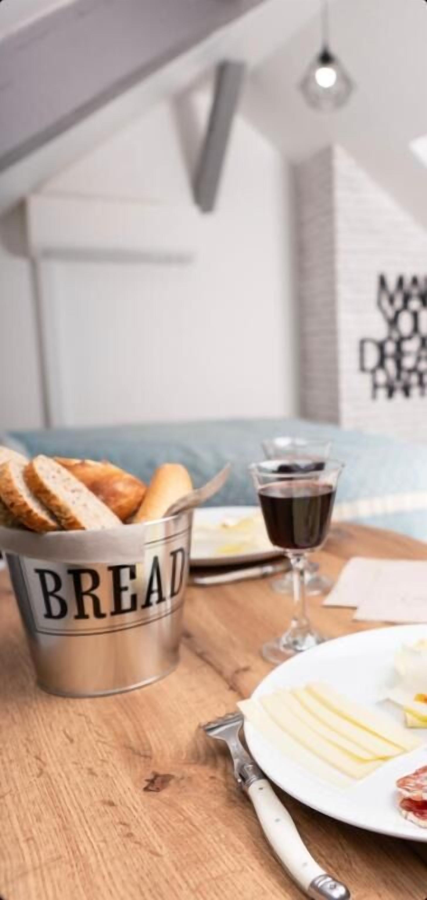 A dining table with a metal bucket labeled "BREAD," a glass of red wine, and a plate with cheese and meat.