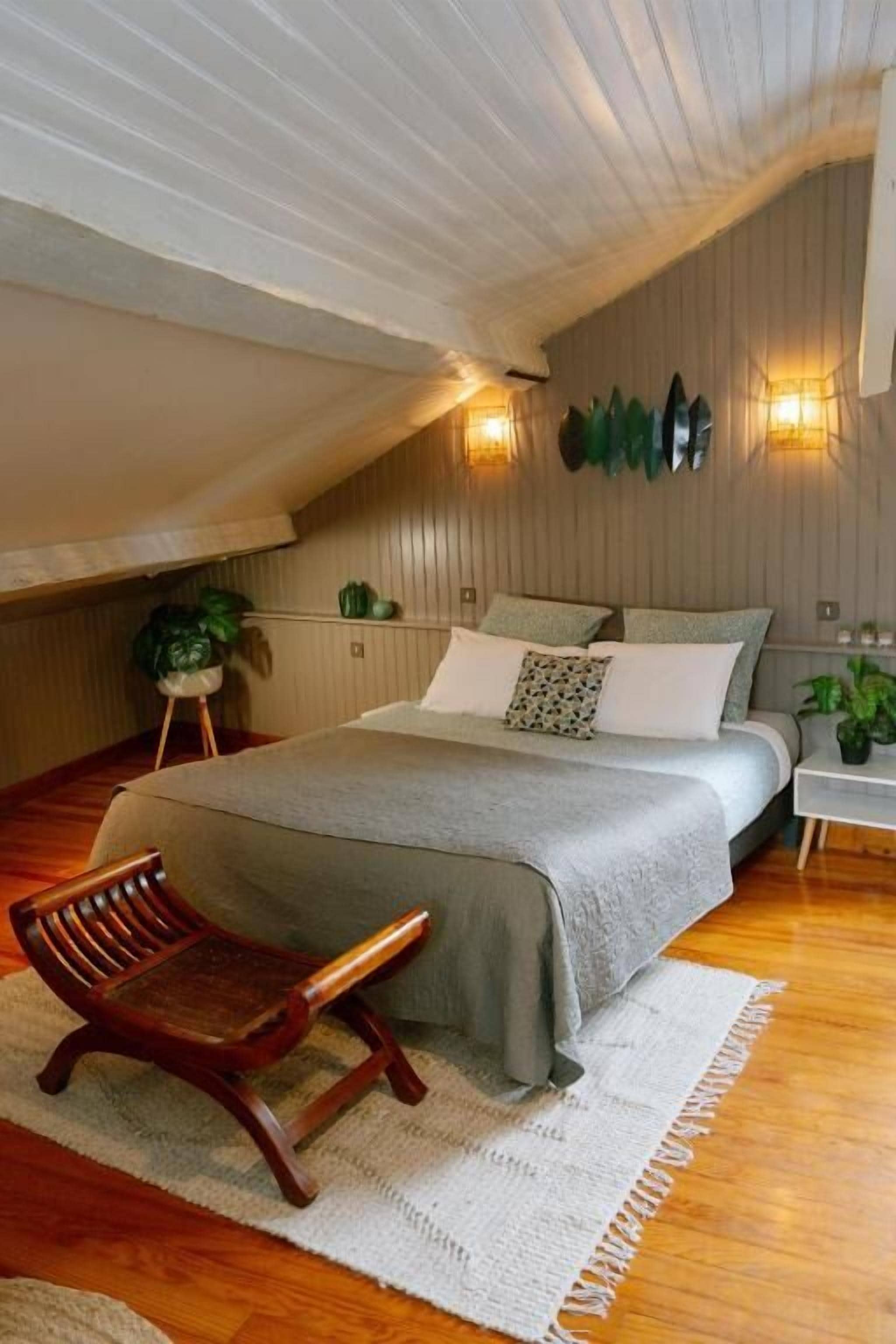 A bedroom with a sloped ceiling, a bed with a grey blanket, a wooden chair, a small table with a plant, and wall-mounted lights.