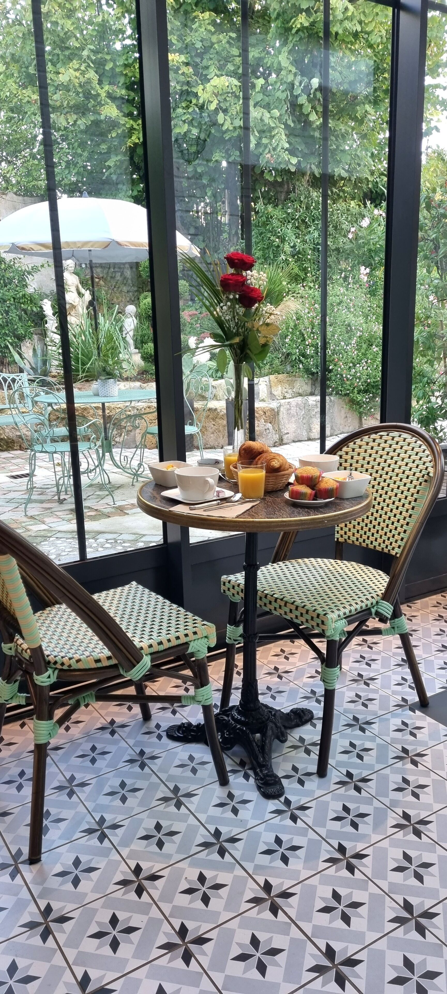 A patio with a round table, wicker chairs, and a view of greenery through large windows.