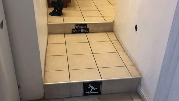 Eine Treppe mit einem Schild, das „Vorsicht, rutschige Stelle“ auffordert, und einem Piktogramm eines laufenden Mannes, das auf Stolpergefahr hinweist.