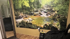 A room with a large nature-themed mural, a wooden floor, a black couch, and a small table with a bag. - The Single Vacation Rental Dwelling (Orlando)