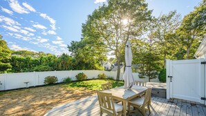 Une terrasse en bois avec une table et des chaises, entourée d’une clôture blanche et d’une végétation luxuriante.