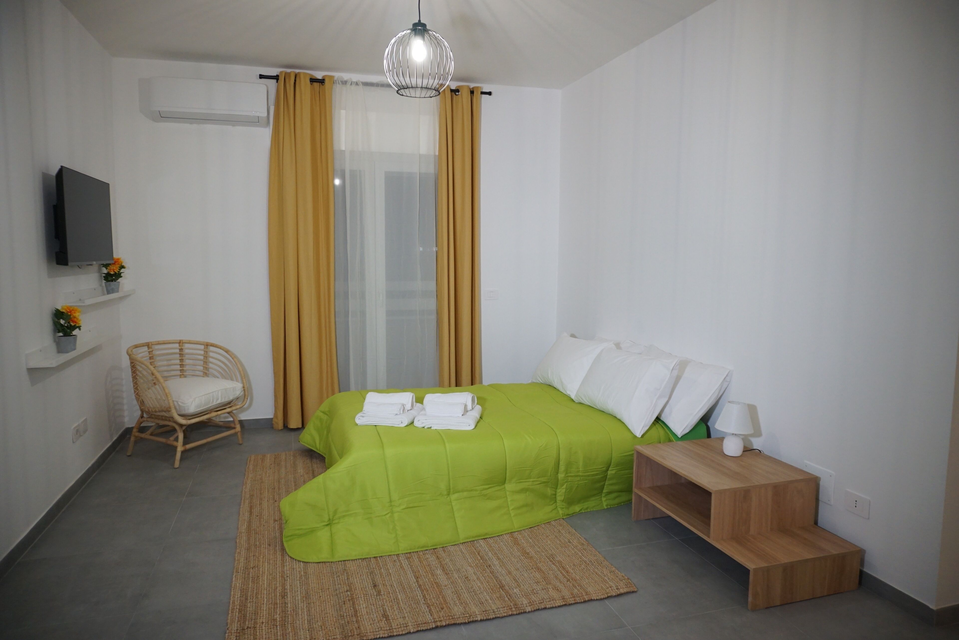 A modern hotel room with a green bed, a wooden nightstand, a wicker chair, and a television on the wall.