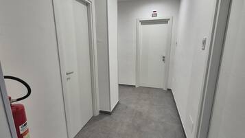 Un pasillo con puertas blancas, un extintor a la izquierda y un detector de incendios a la derecha.