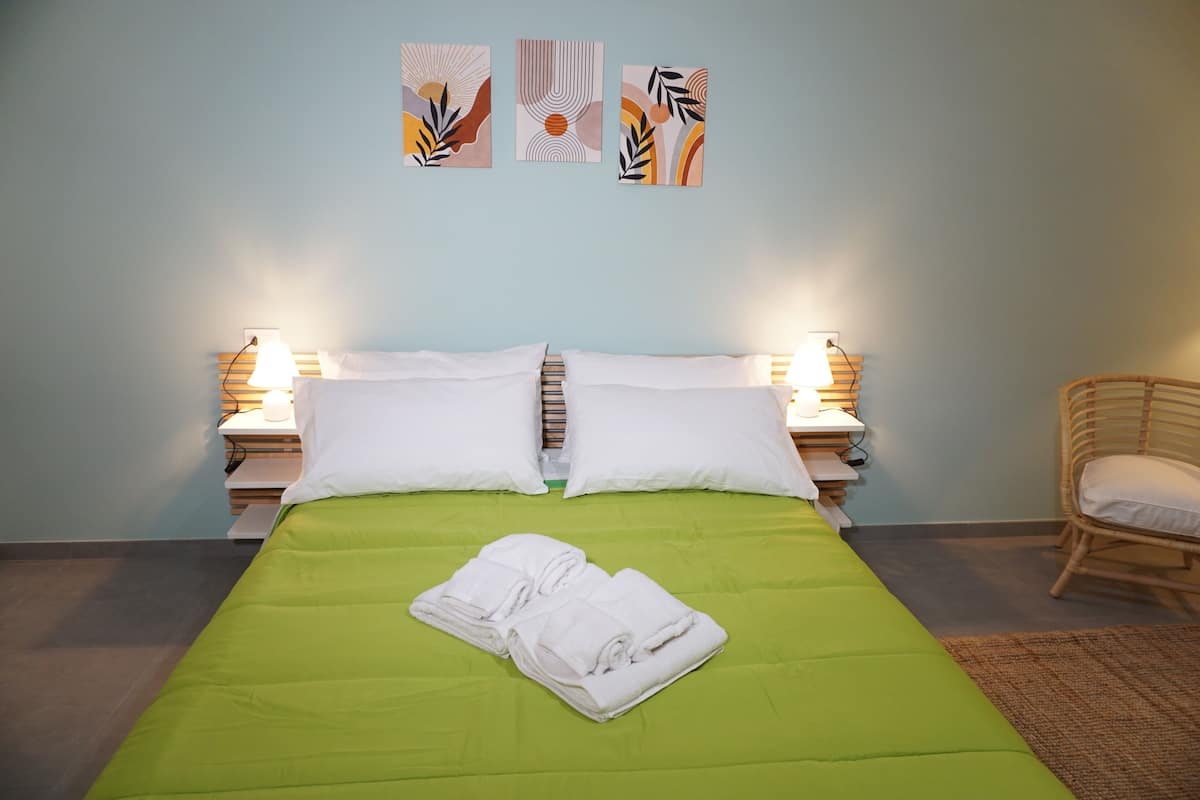 A neatly made bed with white linens and a green bedspread, flanked by two bedside lamps, in a room with light blue walls and abstract artwork above the bed.