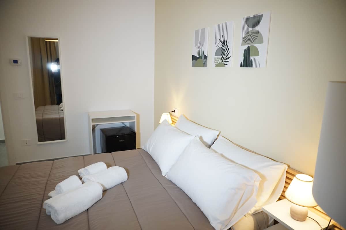 A hotel room with a bed, a bedside table with a lamp, a mirror, and artwork on the wall.