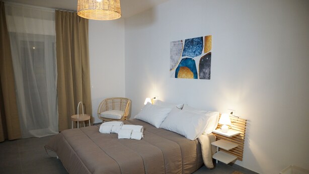 Un lit bien fait, recouvert d’une couverture marron, une chaise en osier, une table de chevet avec une lampe et une œuvre d’art accrochée au mur.
