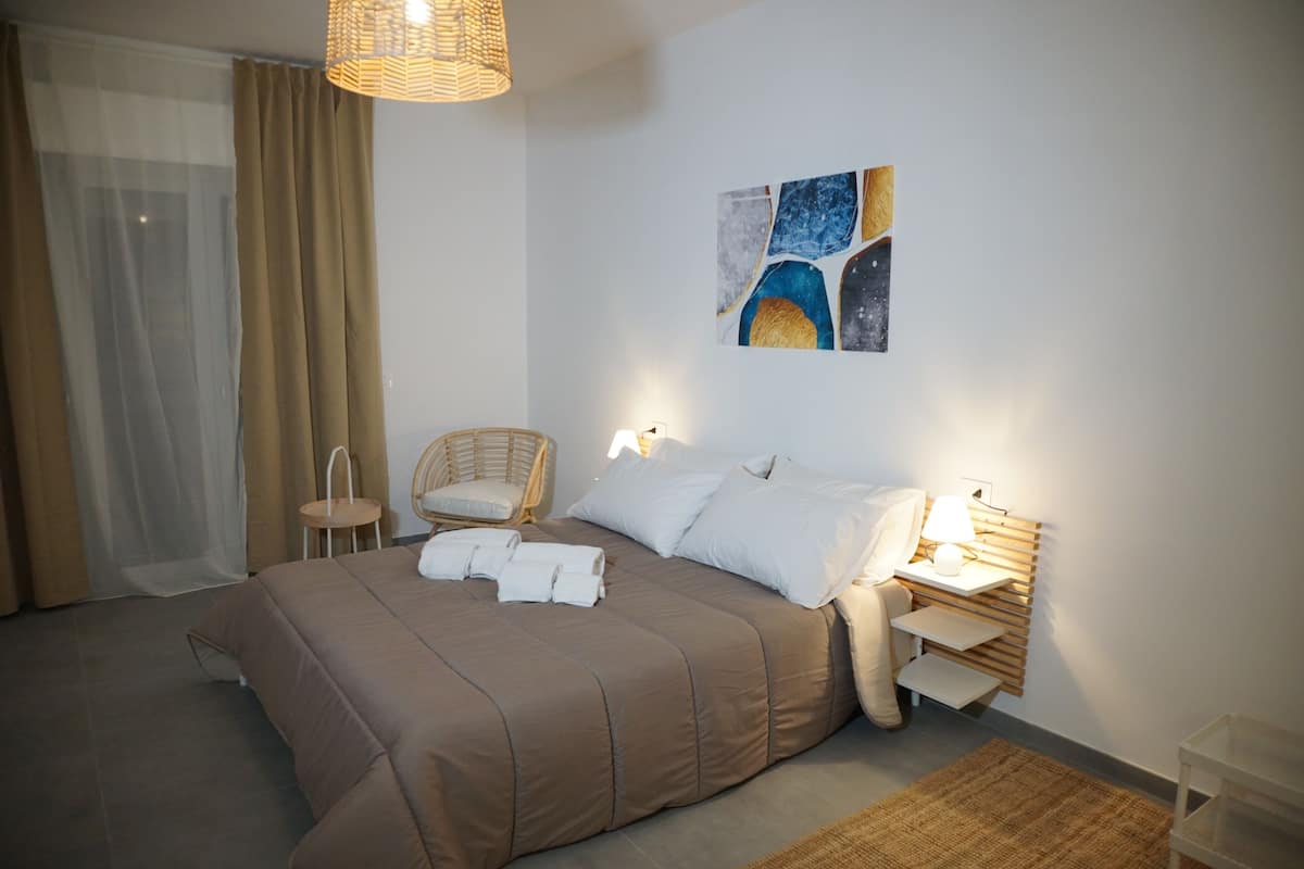 A neatly made bed with a brown bedspread, a wicker chair, a bedside table with a lamp, and a wall art piece.