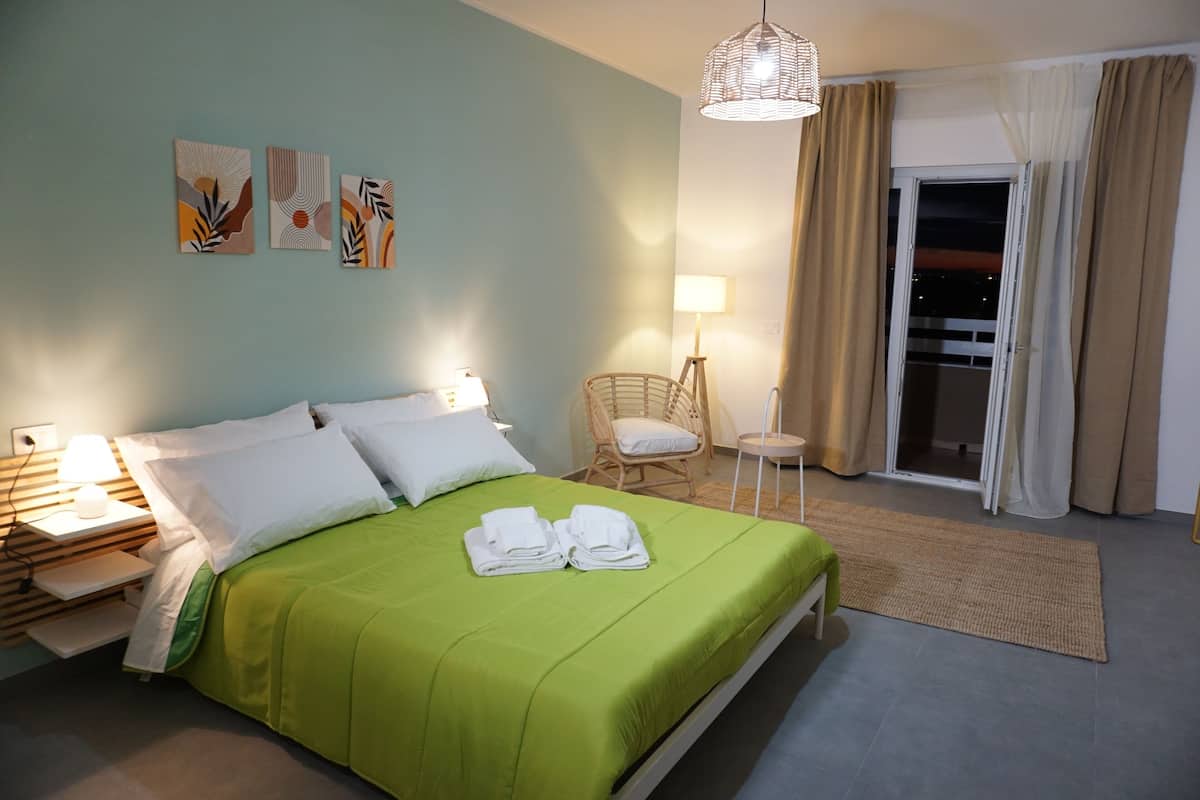 A hotel room with a green bed, white pillows, a wicker chair, a nightstand with a lamp, and artwork on the wall.