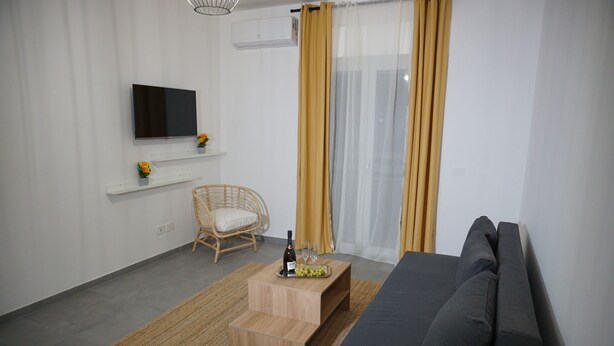 Un salon moderne avec un téléviseur à écran plat, un canapé, un fauteuil en osier, une petite table basse en bois sur laquelle se trouvent une bouteille et des verres, et une fenêtre ornée de rideaux jaunes.