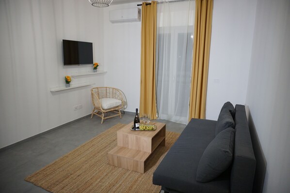 Ein modernes Wohnzimmer mit Flachbildfernseher, einer Couch, einem Korbsessel, einem kleinen Holztisch mit Flasche und Gläsern sowie einem Fenster mit gelben Vorhängen.