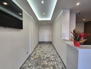 A hallway with a tiled floor, a flat-screen TV mounted on the wall, a reception desk with a potted plant, and a door at the end. - Farina Rooms & Apartments (San Nicola La Strada)