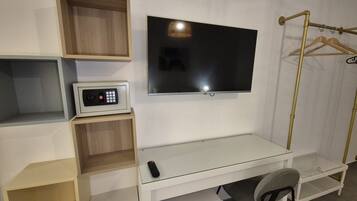 Une chambre compacte équipée d’un téléviseur fixé au mur, d’un four à micro-ondes, d’un petit bureau avec une chaise et d’un porte-vêtements.