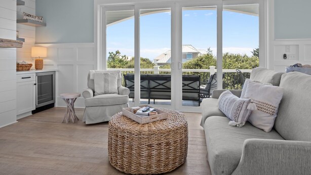 A spacious living area with a large window, a sofa, a chair, and a wicker ottoman. - Silver Lining- Luxury Retreat in Dolphin Ridge (Emerald Isle)