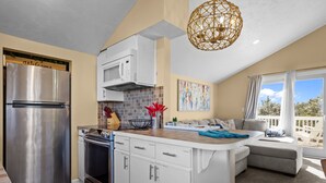A modern kitchen with a stainless steel refrigerator, microwave, and oven. There is a dining area with a sofa and a view of the outdoors. - The Bedloe: Cozy Beach Cottage (Emerald Isle)