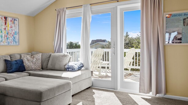 A living room with a sectional sofa, a white ceiling, and a sliding glass door leading to a balcony with white chairs. - The Bedloe: Cozy Beach Cottage (Emerald Isle)