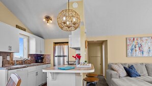 A modern kitchen with white cabinets, a stainless steel refrigerator, and a hanging light fixture. - The Bedloe: Cozy Beach Cottage (Emerald Isle)
