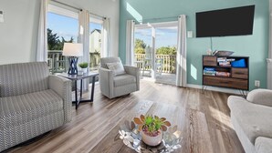 A spacious living room with a wooden ceiling, large windows, a flat-screen TV, and a balcony with a view of the surrounding area. - The Jackson: Cozy Beach Cottage with Pool! (Emerald Isle)