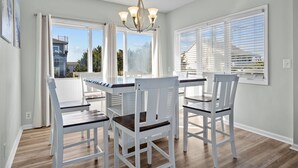 A dining area with a white table and chairs, a chandelier, and large windows with blinds. - The Jackson: Cozy Beach Cottage with Pool! (Emerald Isle)