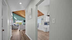 A hallway with wooden flooring, a kitchen with white cabinets, and a living area with a couch and a coffee machine. - The Jackson: Cozy Beach Cottage with Pool! (Emerald Isle)