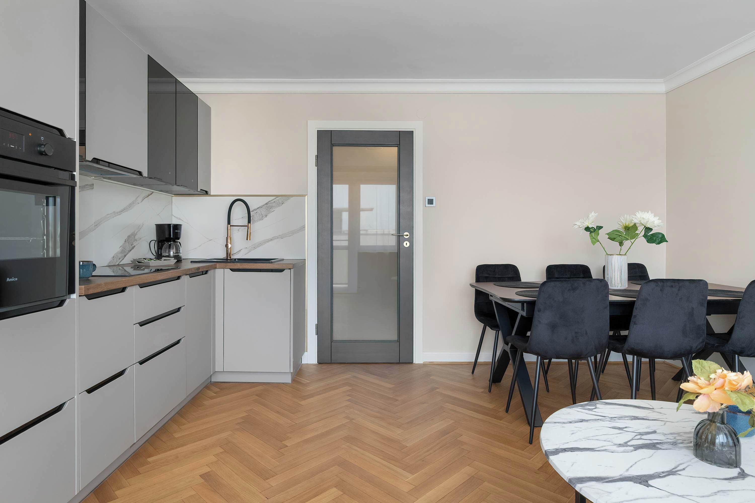 A modern kitchen with grey cabinets, a built-in oven, and a coffee maker. There is a dining area with a table and chairs, and a door leading to another room.