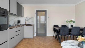 Una cocina moderna con gabinetes grises, horno empotrado y cafetera. Cuenta con un comedor con mesa y sillas, y una puerta que da a otra habitación.