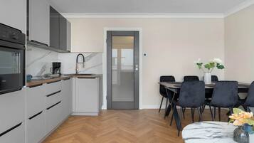 A modern kitchen with grey cabinets, a built-in oven, and a coffee maker. There is a dining area with a table and chairs, and a door leading to another room.