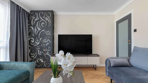 Una sala de estar moderna con un sofá gris, un sofá azul, una mesa de centro con un jarrón blanco y flores blancas, un televisor de pantalla plana sobre un mueble blanco, y una pared con diseño.