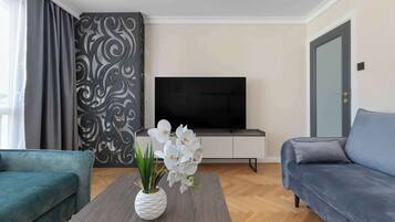 A modern living room with a grey sofa, a blue sofa, a coffee table with a white vase and white flowers, a flat-screen TV on a white cabinet, and a patterned wall.