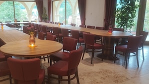 Un vaste espace dédié à la salle à manger, avec de nombreuses tables et chaises, de grandes fenêtres offrant une vue sur la verdure et des plantes en pot.