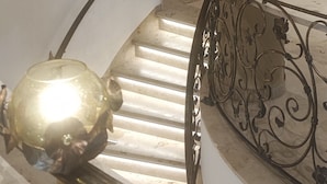 Un escalier en marbre, surmonté d’une sphère dorée, une rampe ornée de motifs floraux et un luminaire placé au-dessus.