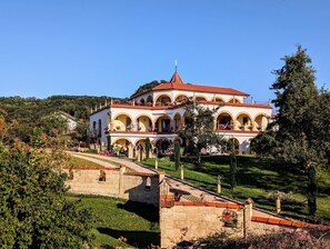 Un edificio grande de varios pisos con techo de tejas rojas, rodeado de abundante vegetación y un puente de piedra.