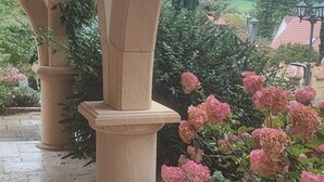 Un espacio al aire libre cubierto con columnas de piedra, piso de baldosas y un jardín de flores rosas.