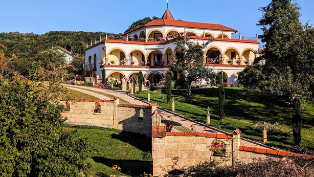 Un edificio grande de varios pisos con techo de tejas rojas, rodeado de abundante vegetación y un puente de piedra.