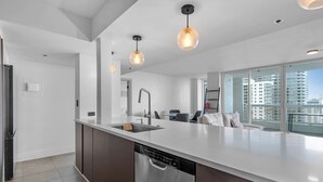 Une cuisine moderne dotée d’un grand îlot central, d’un lave-vaisselle en acier inoxydable et d’un évier encastré. Des luminaires suspendus sont placés au-dessus de l’îlot, et de grandes fenêtres offrent une vue sur la ville.