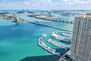 An aerial view of a marina with numerous boats docked, a bridge connecting the islands, and high-rise buildings in the background.